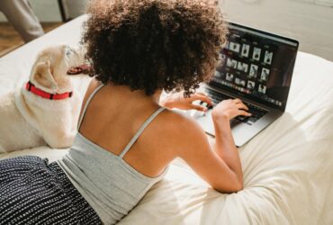 black woman browsing laptop with dog on bed